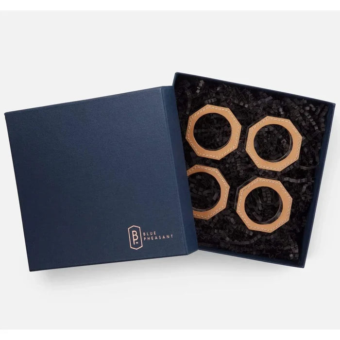 A navy blue gift box with Blue Pheasant branding opens to reveal four Arthur Leather Camel Napkin Rings, stylishly arranged on black shredded paper—an elegant addition to any table setting.