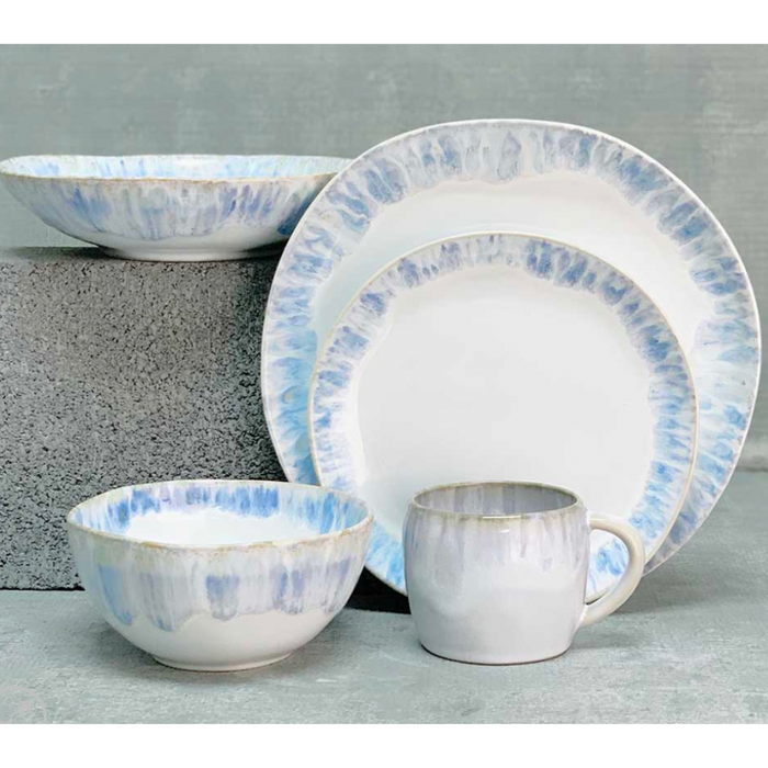 The Costa Nova Brisa Blue Dinnerware set features two plates, a bowl, and a mug in white with blue and tan watercolor edges, displayed on a gray surface. This stoneware set is both elegant and versatile.