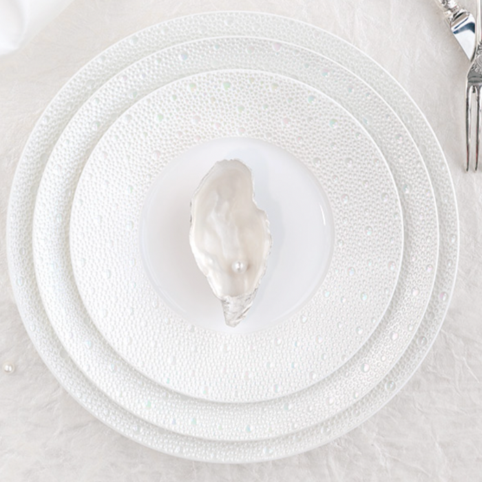 A white oyster shell with a pearl sits on a Bernardaud Ecume Dinnerware Perle plate, layered over two larger textured plates, with a fork and knife at the top right, all set against a pristine white background.