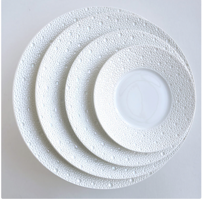 Four white Ecume Dinnerware Perle plates by Bernardaud are stacked on a flat, light surface. The rims have a raised pebble-like texture and subtle mother-of-pearl finish, while the centers are smooth.