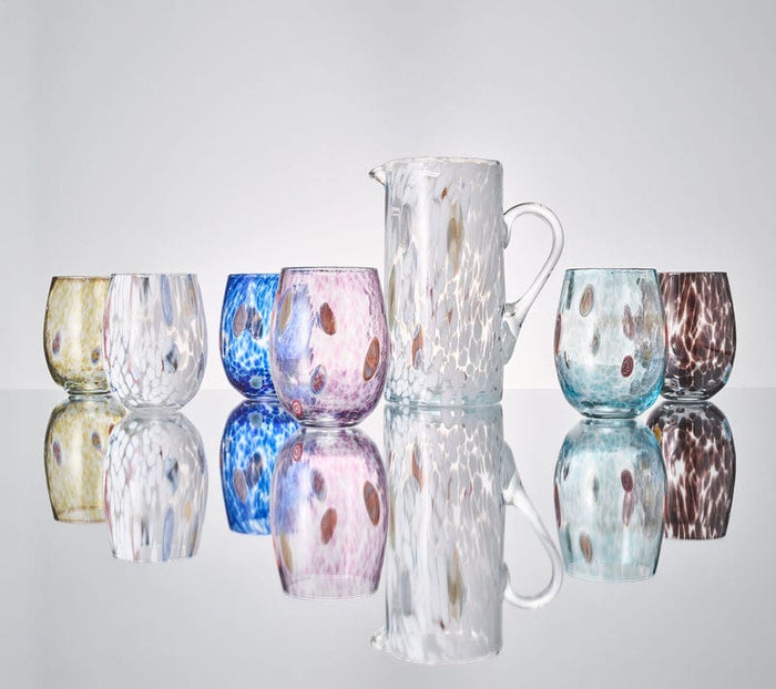 The Kim Seybert Gala Pitcher White and six colorful glass tumblers, crafted in Murano Glass style, are displayed on a reflective surface, creating mirrored reflections below them.
