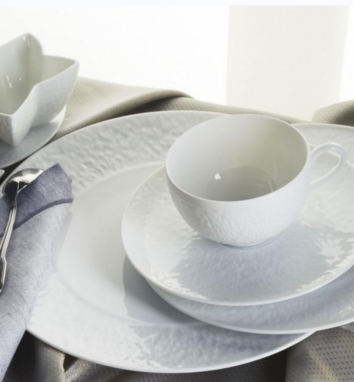 A close-up of a table set with Raynaud Mineral Dinnerware Glazed White—including cup, saucer, bowl, and plates—on a gray napkin and beige tablecloth next to silver cutlery.