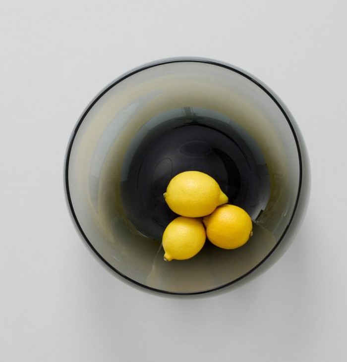 A top-down view of three yellow lemons in the Gary Bodker Nesting Glass Bowl Charcoal, arranged on a light gray surface to showcase the bowl’s handcrafted elegance.